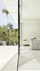Modern minimalist living room with concrete flooring, white walls, and large windows overlooking palm trees.