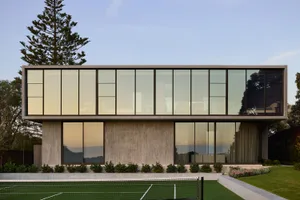 A modern residential building with a concrete exterior, large glass windows, and a tennis court in the foreground.