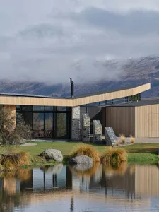 A modern house with a wooden exterior sits on the shore of a calm lake with mountains in the background.