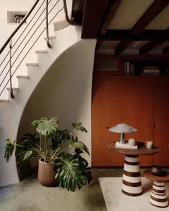 A modern interior design featuring a wooden table with concrete base, potted plants, and a lamp, alongside a curved staircase
