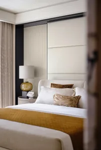 A modern minimalist bedroom featuring a wooden floor, white and gold bedding, and a white lamp with a gold base.