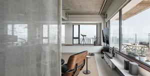 A modern, minimalist living room with concrete finishes, a large window, and a single armchair.