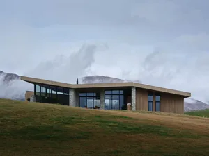 A modern residential building with a wooden exterior and concrete accents, situated on a grassy hilltop.