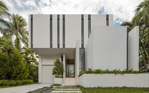 A modern house with white concrete walls and large windows surrounded by green palm trees.