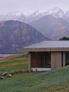 Modern minimalist residential exterior featuring a wooden façade with a metal roof, set against a backdrop of majestic snowy 