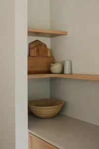 A minimalist kitchen with wooden shelves holding cutting boards, a jug, a small vase, and a woven basket.