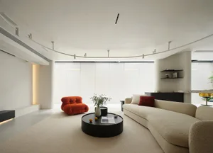 A modern minimalist living room with neutral tones, featuring a beige sofa, an orange accent chair, and a black round coffee 