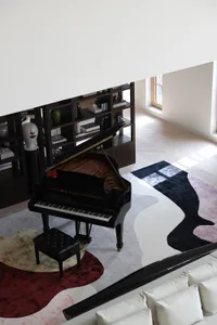 A modern living room featuring a black grand piano on a colorful abstract rug, with white cushions on a sofa and a bookshelf 
