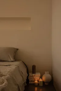 A minimalist bedroom with a bed covered in beige textiles, a stack of books, candles, and a ceramic vase on a bedside table.