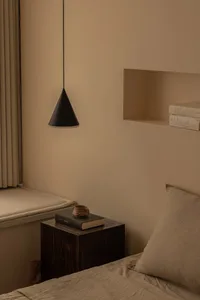 A minimalist bedroom featuring a wooden side table with a book and a ceramic mug, next to a window with beige curtains and a 