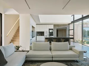 Modern minimalist living room with white furniture and light wood accents, featuring a seating area, staircase, and kitchen.
