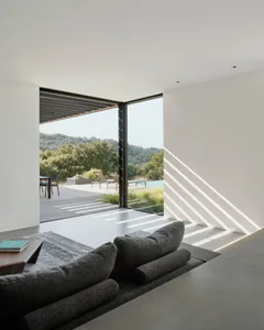 A minimalist living room with a concrete floor, dark furniture, and a large window showcasing a natural landscape.