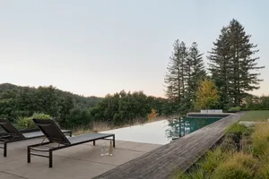 A minimalistic outdoor space featuring a wooden platform with lounge chairs and a swimming pool surrounded by tall trees.