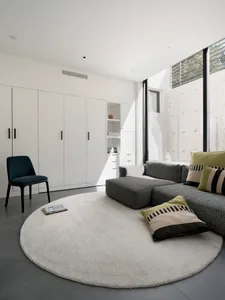 A minimalist living room with a circular white rug, a grey sofa with green and striped cushions, a grey ottoman, and a concre