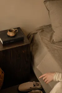 A minimalist bedroom with light-colored bedding, a wooden nightstand, and a black book with a ceramic cup on top.