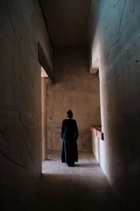 A woman standing in a dimly lit corridor with concrete walls and a wooden beam. She is wearing a black outfit.