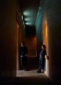 Two individuals standing in a dimly lit, minimalist concrete hallway with rectangular openings in the ceiling.
