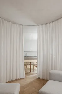 A spacious residence with white curtains, wooden flooring and furniture, and a view into the kitchen area.