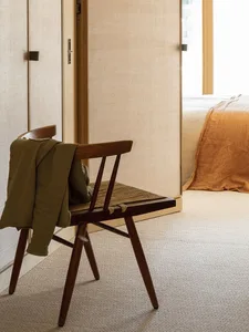 Minimalist room featuring an empty wooden chair in front of a door and a bed with an orange blanket visible in the background