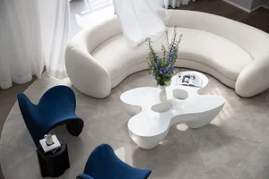 A modern interior design featuring a white circular sofa, blue armchairs, and a glass coffee table with a vase of purple flow