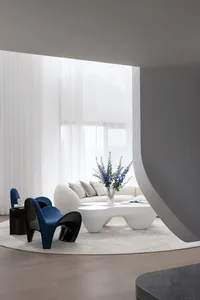 A sleek, modern living room featuring white curtains, a white sofa, and a unique white coffee table with a vase of blue flowe