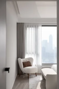 A minimalist apartment features a modern, white chair with a textured pillow, a white sofa, and grey curtains by a large wind