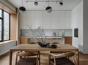 Modern minimalist kitchen with white cabinetry, wooden table and chairs, and a white backsplash with a decorative plant arran