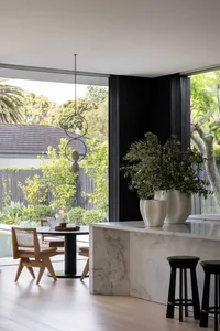 Modern kitchen with marble countertops, concrete flooring, and wooden dining chairs near a large window.