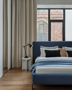 A minimalist bedroom with a blue upholstered bed, beige curtains, and a wooden floor.