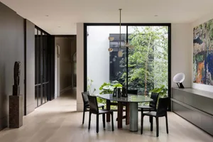 A minimalist dining room featuring a sleek black table with four chairs, large windows overlooking greenery, and modern light