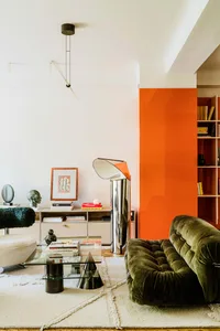 A modern living room with a green sofa, glass coffee table, and an orange accent wall