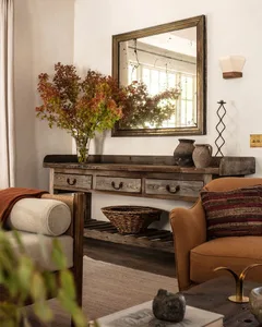 A rustic living room with a wooden console table, orange armchair, and a large mirror reflecting plants.