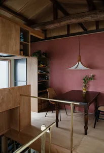 A minimalist kitchen with wooden cabinets and flooring, featuring a wooden table and chairs, and a pink wall.