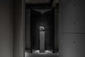 A minimalist exhibition space featuring a metal monolith against a dark background.