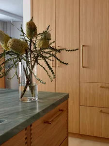 Modern minimalist kitchen with wooden cabinets and marble countertop featuring Banksia flowers in a vase.