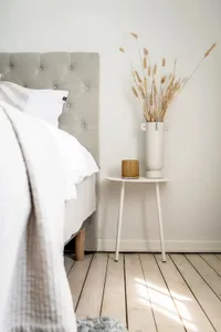 A minimalist bedroom with a fabric bed against a white wall, a white side table with a vase of dried plants and a candle hold