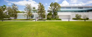 A modern residential home featuring a large concrete driveway, modern glass walls, and lush greenery.