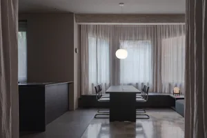A minimalist residential interior featuring light wood and cement surfaces, with a large window letting in natural light.