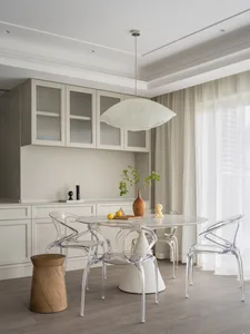 Modern minimalist kitchen with marble countertops and dining table, featuring transparent chairs and light neutral curtains.