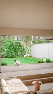 A modern minimalist living room with a white couch, wooden flooring, concrete walls, and lush greenery.