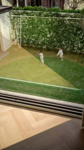 Two children playing on a green lawn surrounded by a glass wall with plants.
