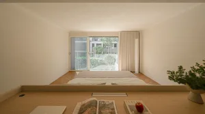 A minimalist bedroom with a low wooden frame bed, white bedding, and a window overlooking trees and buildings.