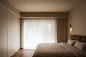 A minimalist bedroom featuring a bed with white bedding, a wall-mounted light fixture, and white curtains