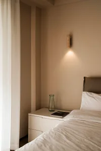 A minimalist bedroom with neutral tones featuring a bed with white bedding, a nightstand with a vase, and a wall-mounted ligh