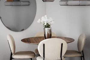 A modern dining room with wooden table, upholstered chairs, metallic accents, and a vase of white flowers.