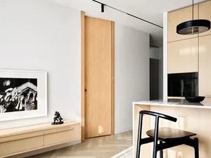 Modern, minimalist kitchen featuring wooden cabinets and flooring, with black pendant lighting and a single black chair.