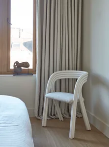 A minimalist bedroom featuring a white plastic chair and a bed with white bedding, with a curtain and artwork on the wall, an
