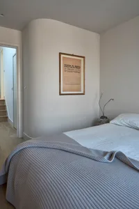 A minimalist bedroom with a light gray blanket on the bed, framed artwork on the wall, and modern lighting.