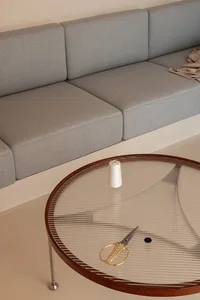 A modern glass coffee table with a wooden frame and metal legs, featuring a spool of white thread, a pair of scissors, and a