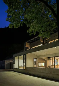 Modern house with concrete walls and glass windows illuminated at night, surrounded by trees.